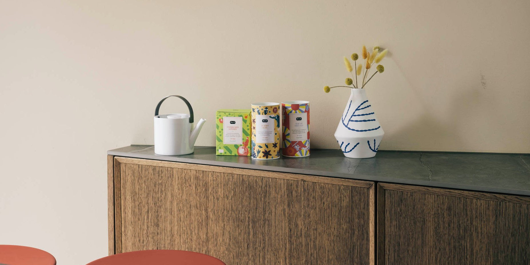 Holzschrank mit dunkler Oberfläche, darauf eine weiße Gießkanne, drei farbenfrohe Teeboxen mit unterschiedlichen Designs und eine weiß-blau gestreifte Vase mit gelben Blumen
