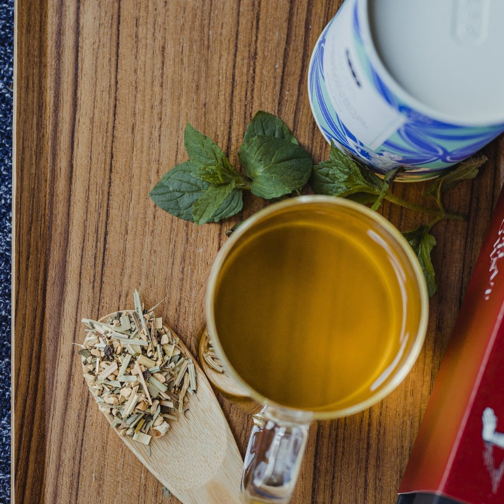 Klare Glastasse mit bernsteinfarbenem Tee auf Holzuntergrund, daneben Holzlöffel mit Kräuterteeblättern und Minze.