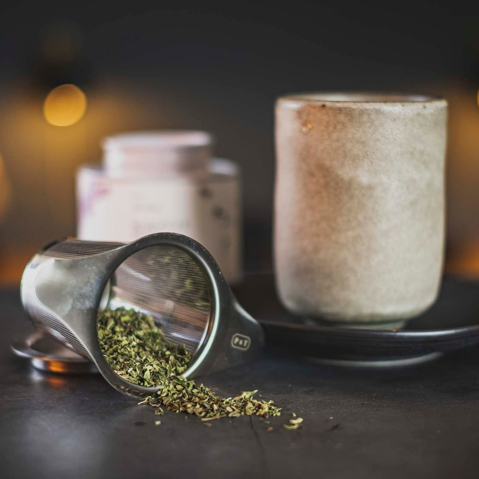 Metall-Teesieb mit grünen Teeblättern neben Keramiktasse auf dunklem Untersetzer