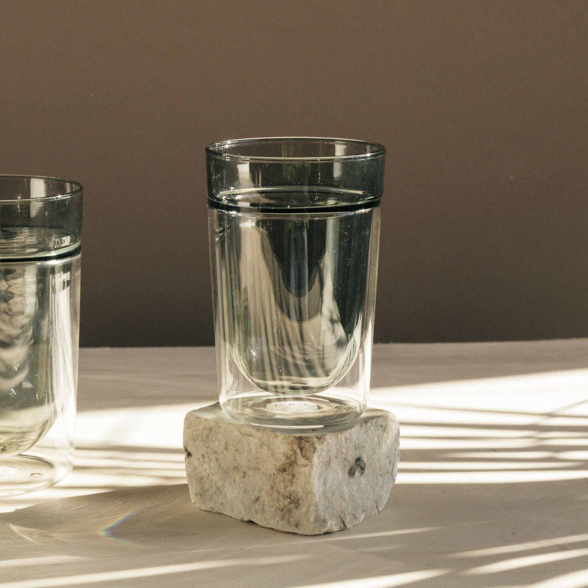 Klares Glas mit Wasser auf einem hellen Steinblock vor braunem Hintergrund.