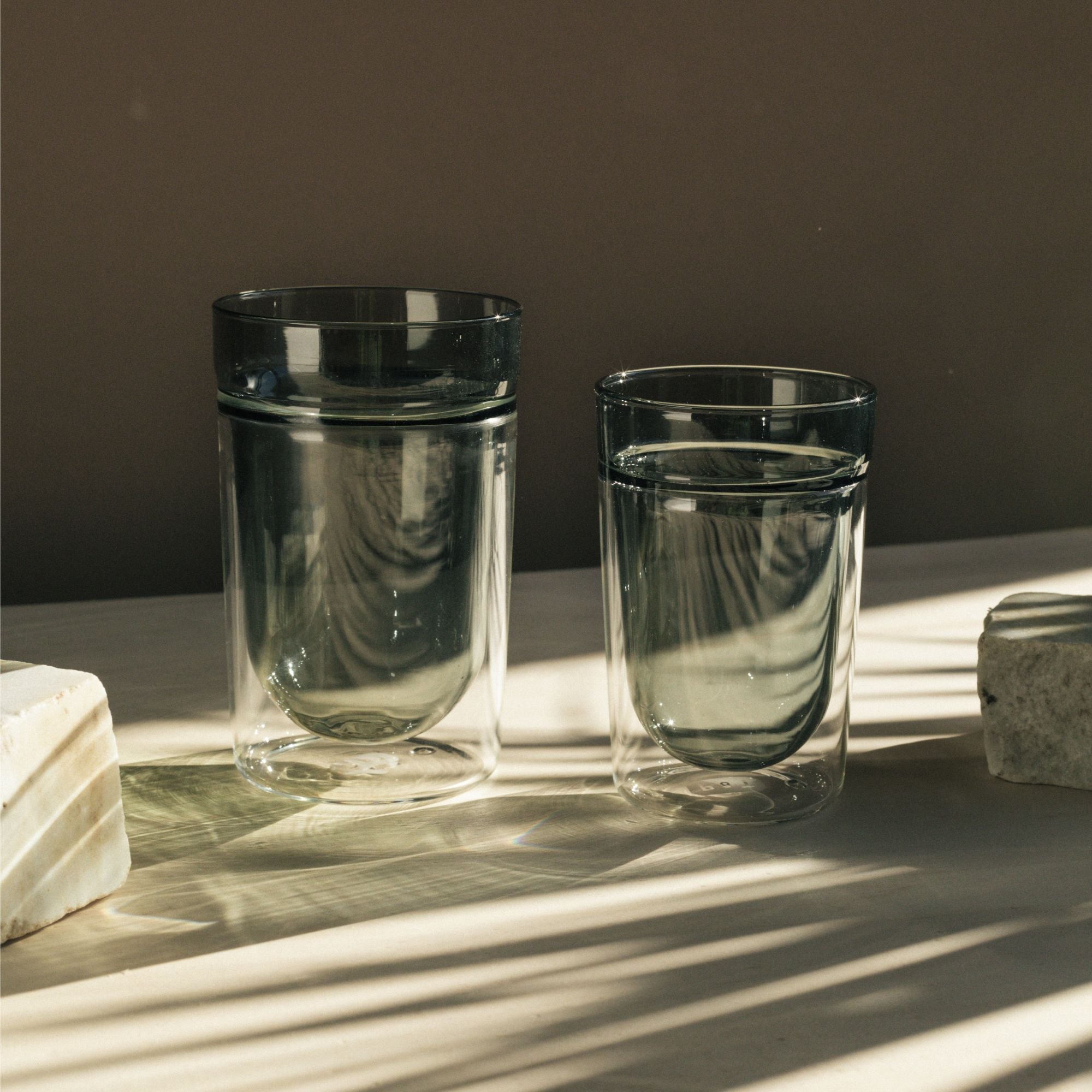 Zwei doppelwandige Gläser mit Wasser auf cremefarbenem Untergrund, umgeben von Natursteinblöcken im Tageslicht.