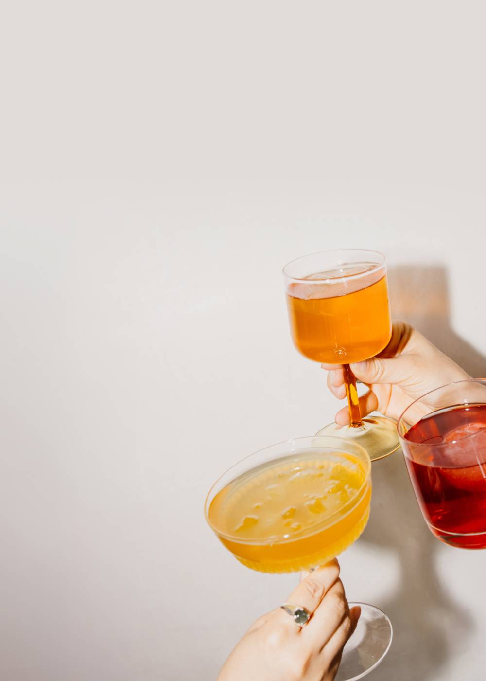 Zwei Hände heben drei Cocktailgläser mit Getränken in Bernstein, Gelb und Rubinrot zum Anstoßen.