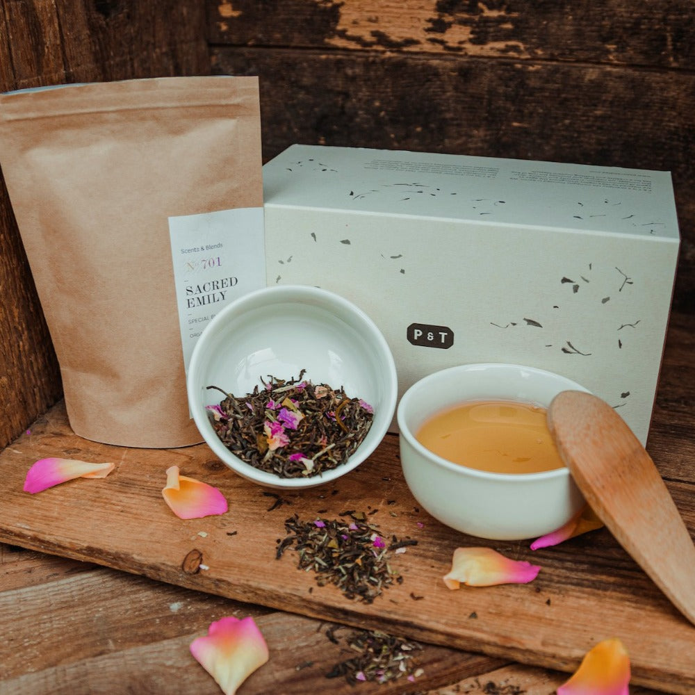 Rustikale Anordnung mit Kraftpapier-Teeverpackung 'Scents & Blends N 701 SACRED EMILY SPECIAL', weißer Box 'P & T', Keramikschalen mit losem Tee und gebrühtem Tee auf Holz mit Blütenblättern.