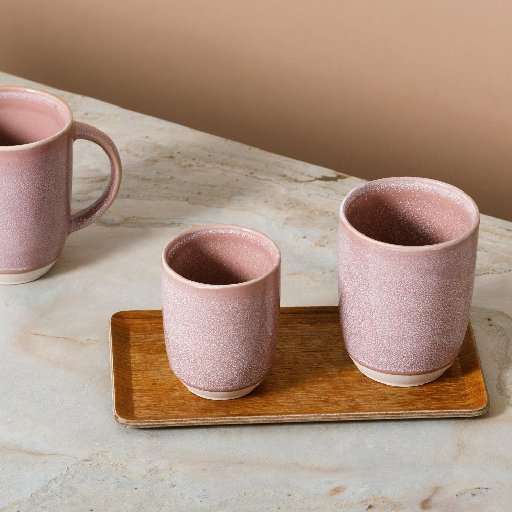 Rosa Keramikbecher-Set mit Speckle-Muster auf Marmor, zwei auf Holztablett, eine Tasse mit Henkel daneben.