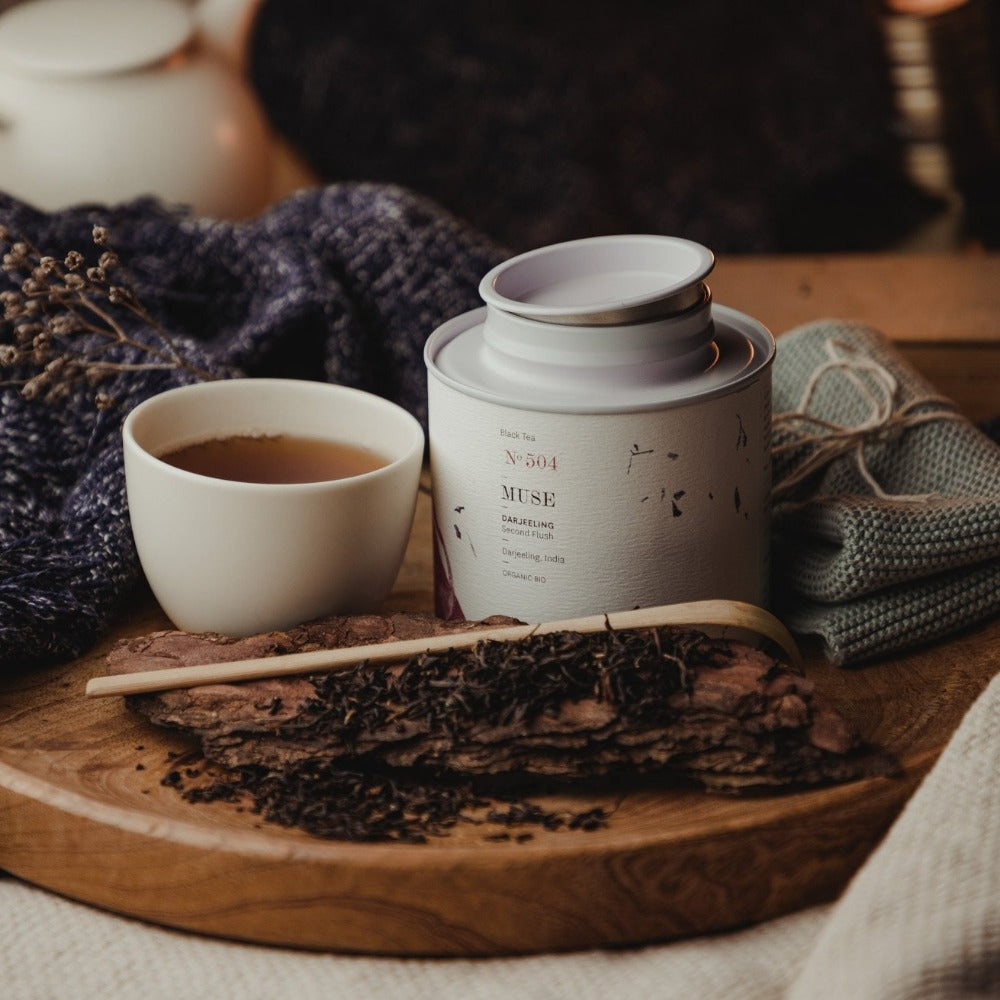 Weißer Keramikbecher mit Darjeeling-Schwarztee, MUSE Nº 504 Dose, lose Teeblätter und Strickstoffe im Hintergrund.