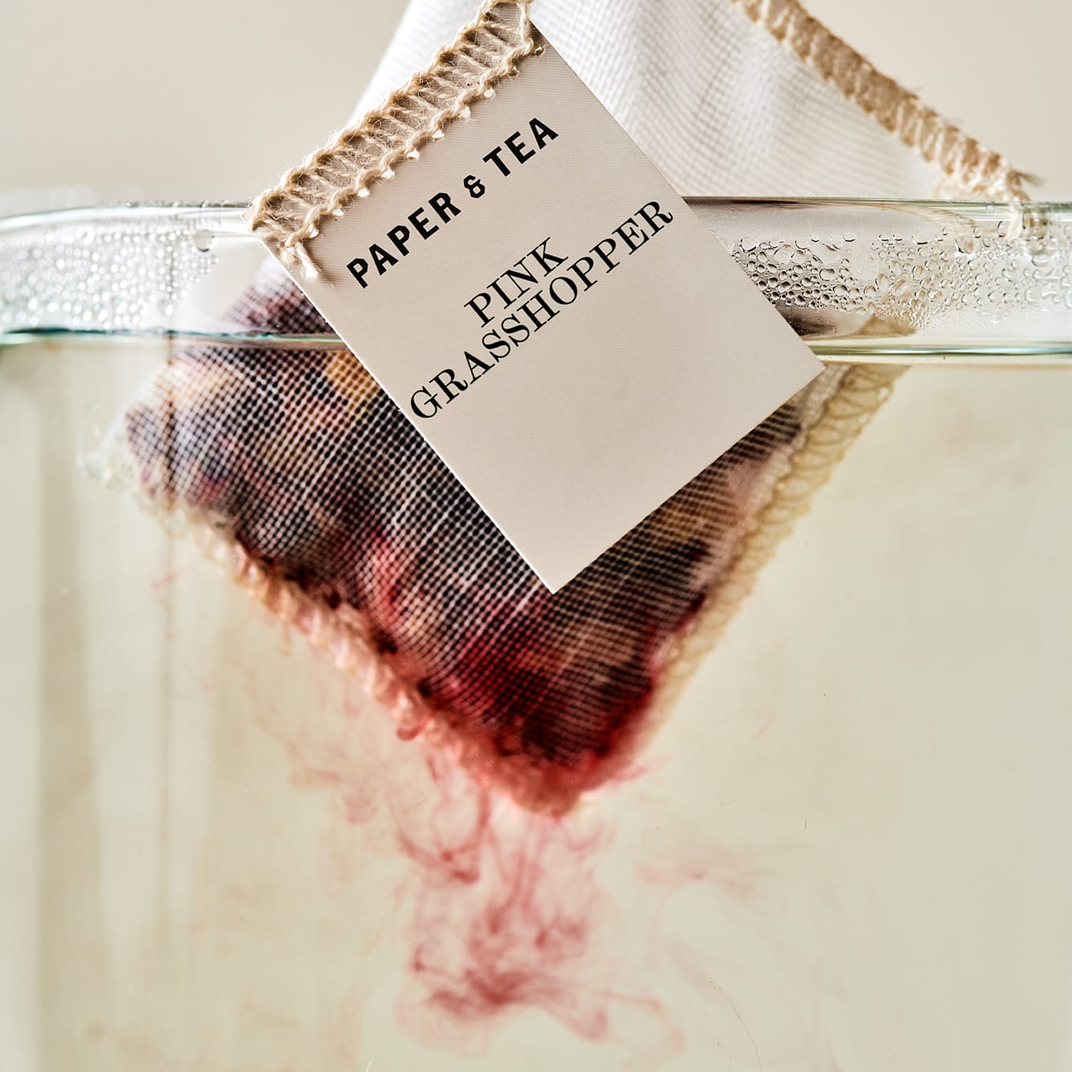 Nahaufnahme eines Teebeutels mit der Aufschrift „PAPER & TEA PINK GRASSHOPPER“, rosa Farbe löst sich im Wasser.