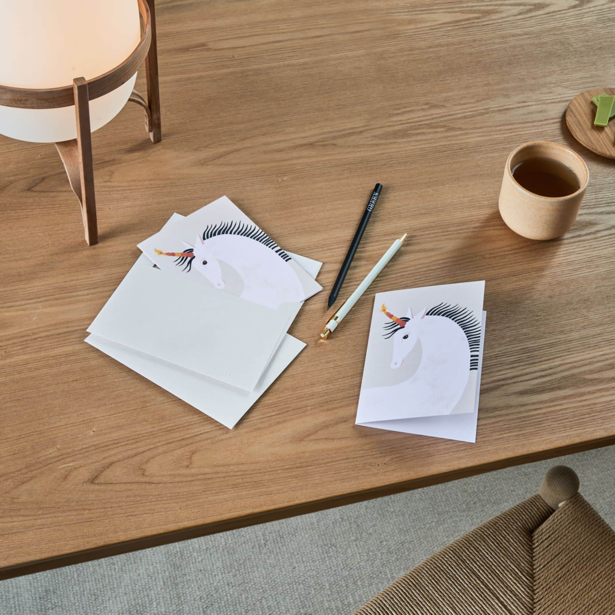 Holztisch mit Einhorn-Karten, schwarzem Bleistift 'GREEN', weißem Stift, Teetasse und Holztablett.