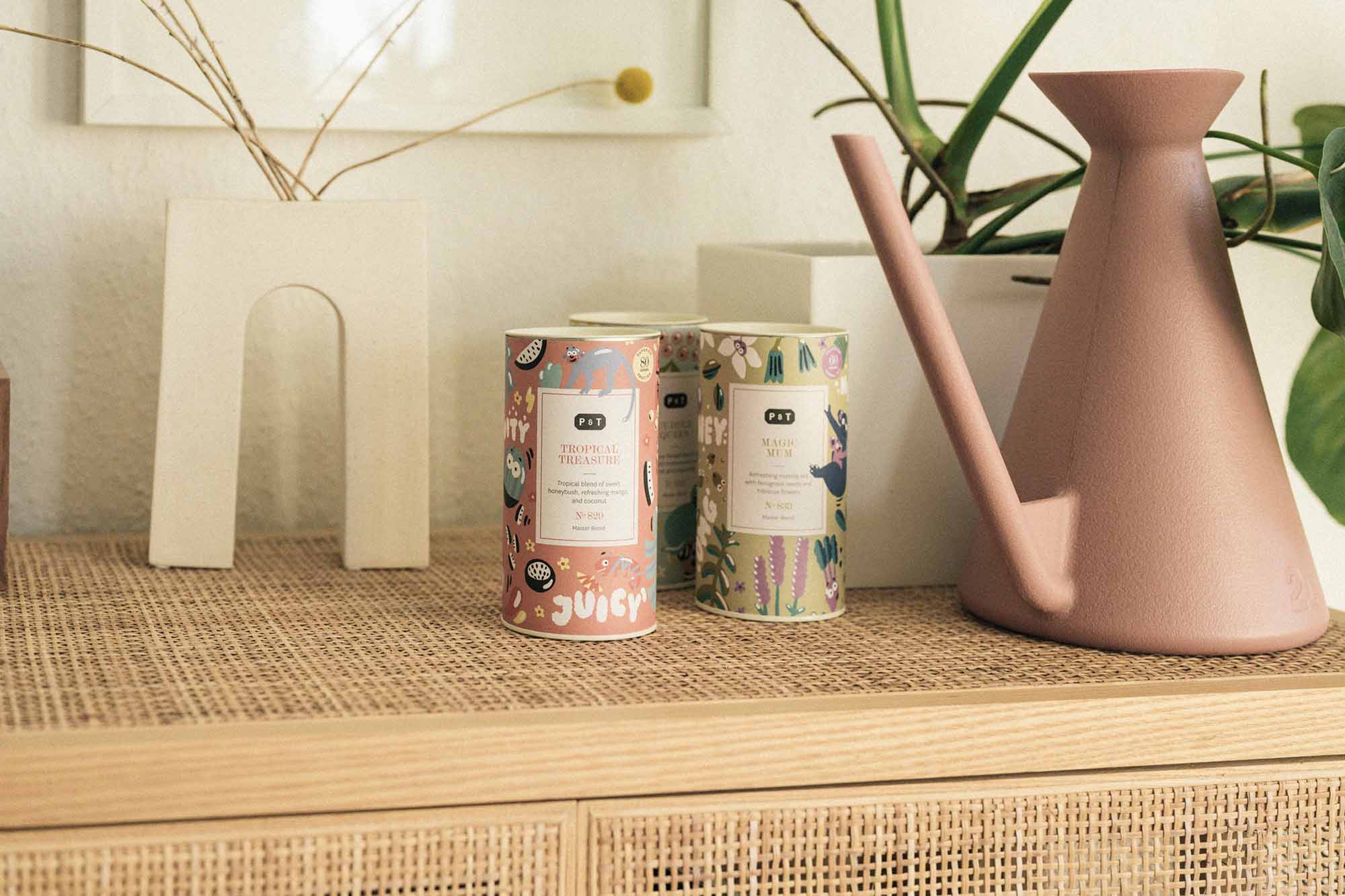 Zwei bunte Teedosen mit Illustrationen, MAGIC MUM und TROPICAL TREASURE, auf Regal neben rosa Gießkanne.