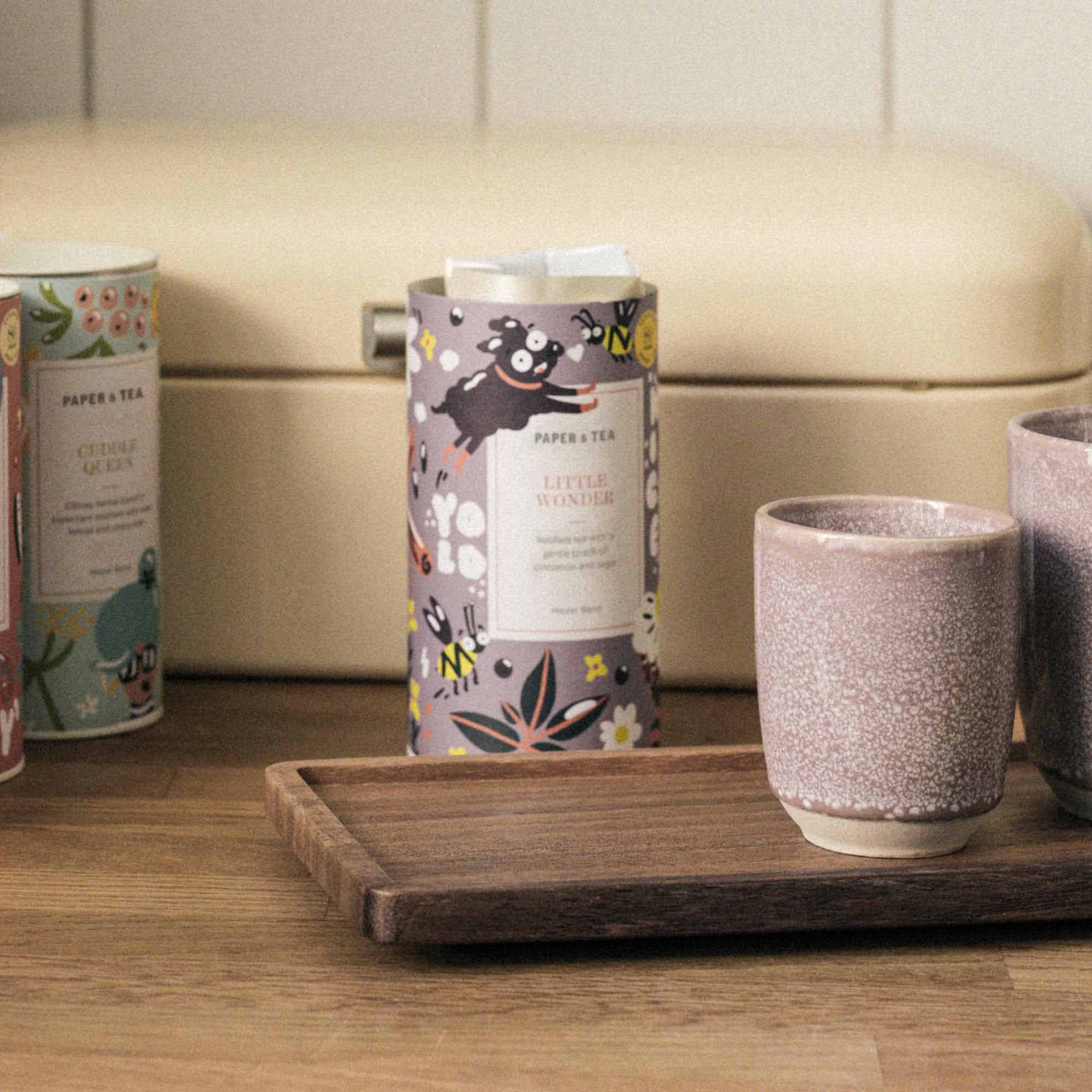Lila Teedose PAPER & TEA LITTLE WONDER mit Comic-Hund und Bienen, Keramikbecher auf Tablett in Küche.
