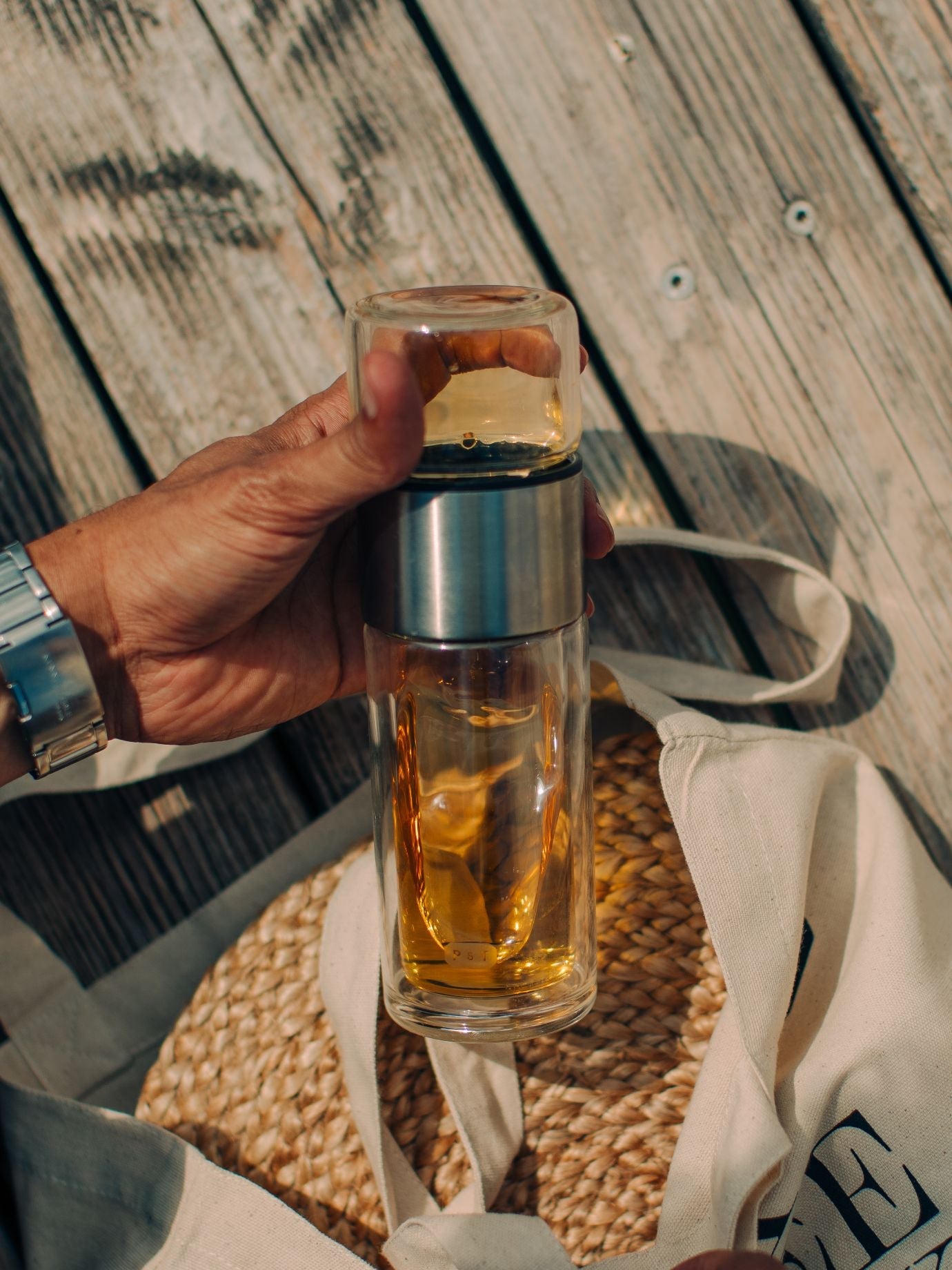 Hand mit silberner Uhr hält Glasbehälter mit gelber Flüssigkeit über beigem Stoffbeutel auf Holzplanken.