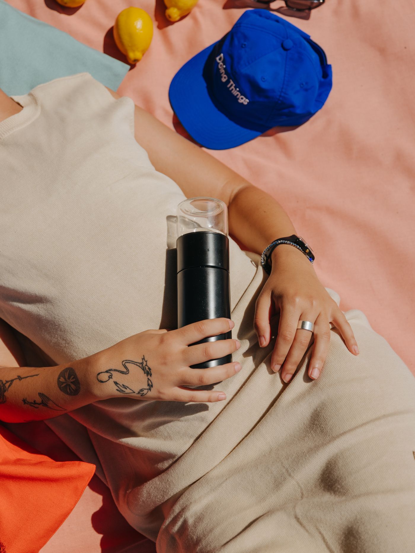 Person liegt auf pfirsichfarbenem Untergrund, hält schwarzen Behälter, blaue 'Doing Things' Kappe, Zitronen und Sonnenbrille daneben.