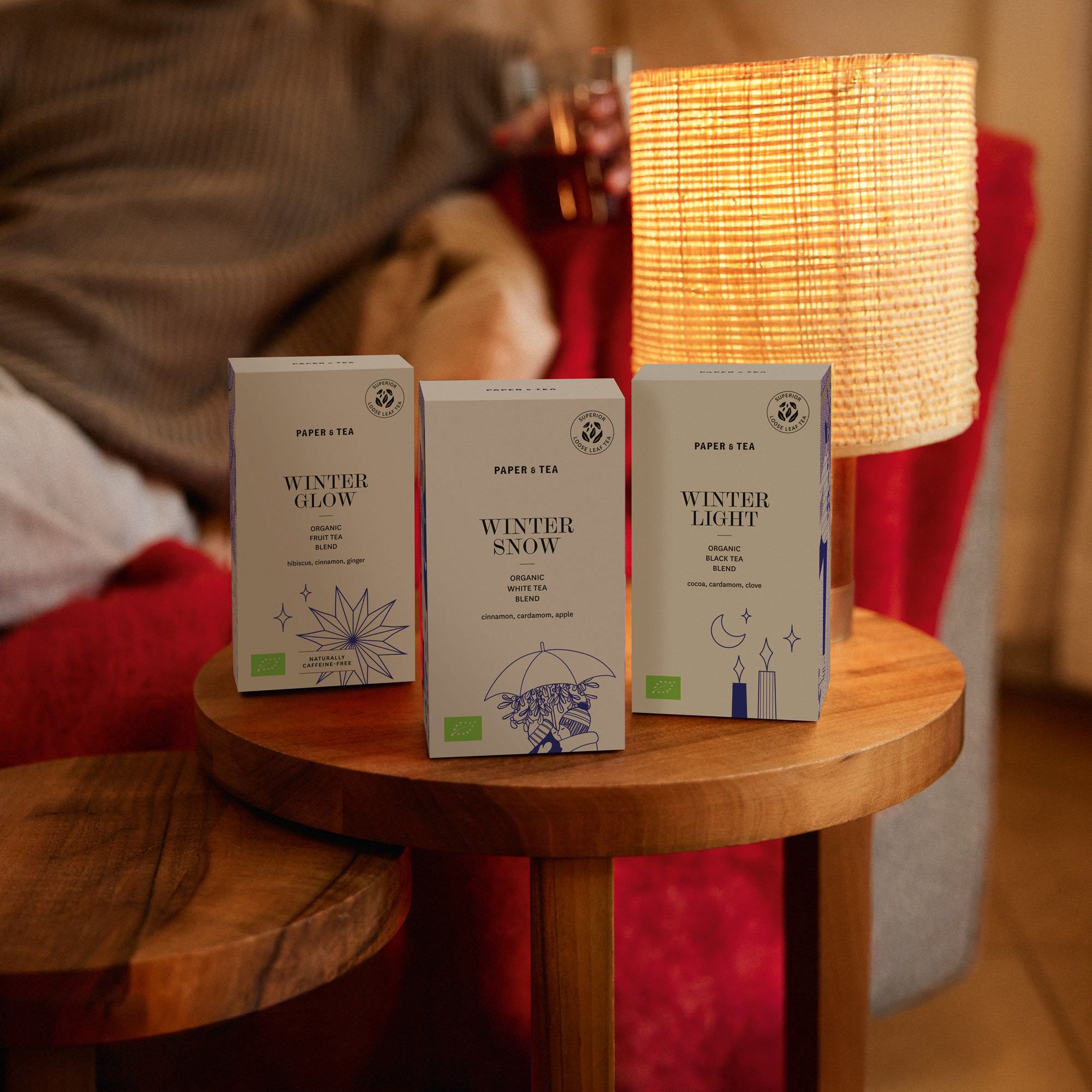 Drei Packungen Paper & Tea Wintertees auf Holztisch mit Lampe und gemütlicher Atmosphäre.