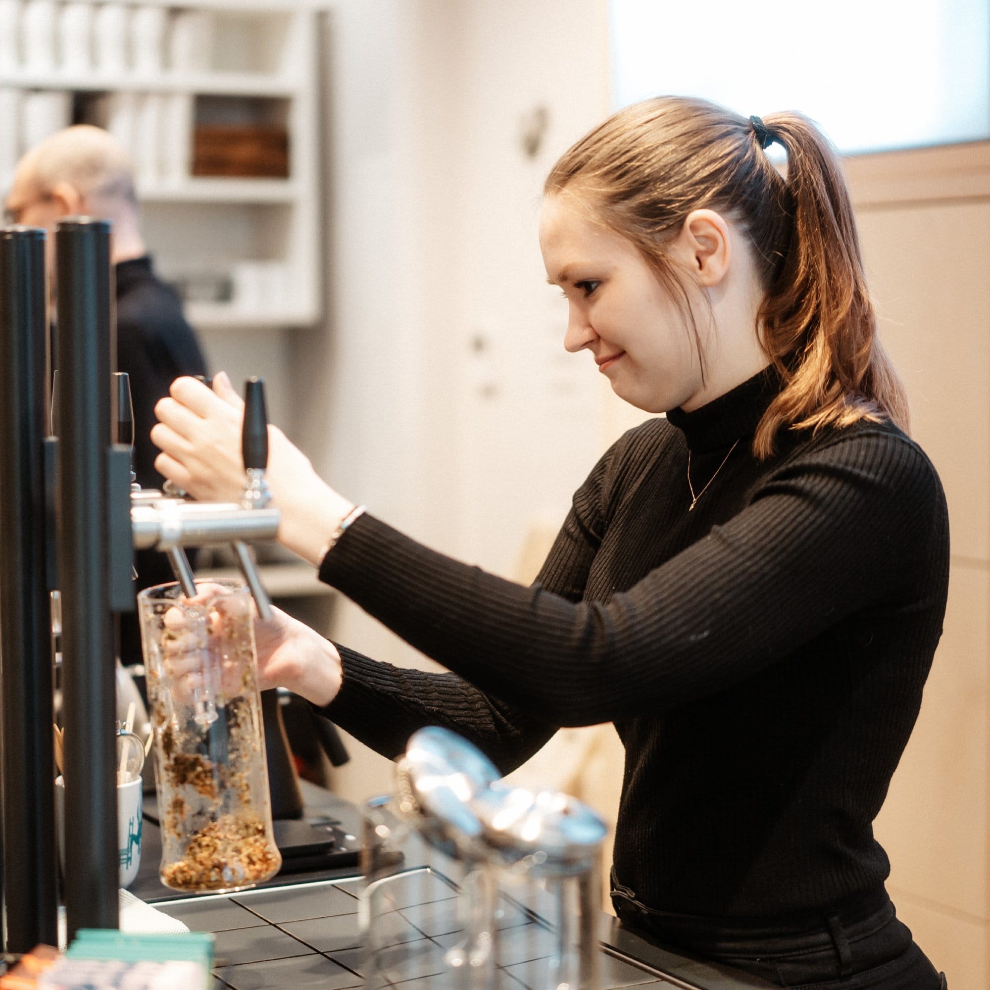 Frau mit Pferdeschwanz im schwarzen Rollkragenpullover füllt Glas im modernen Café aus schwarzem Zapfhahn