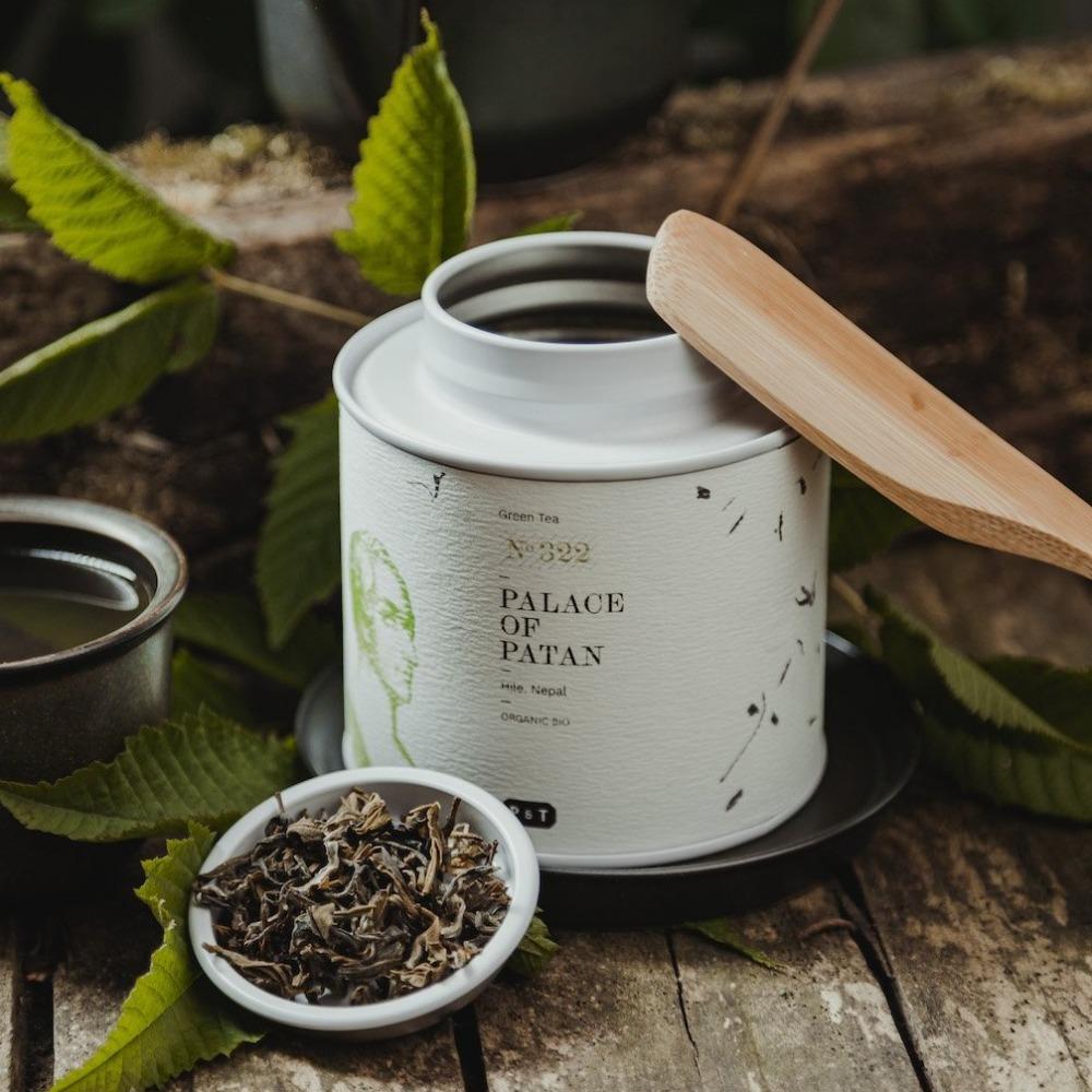 Weißer Teebehälter mit grünem Blattmotiv, getrocknete Teeblätter und frisch gebrühter Tee auf Holztisch.