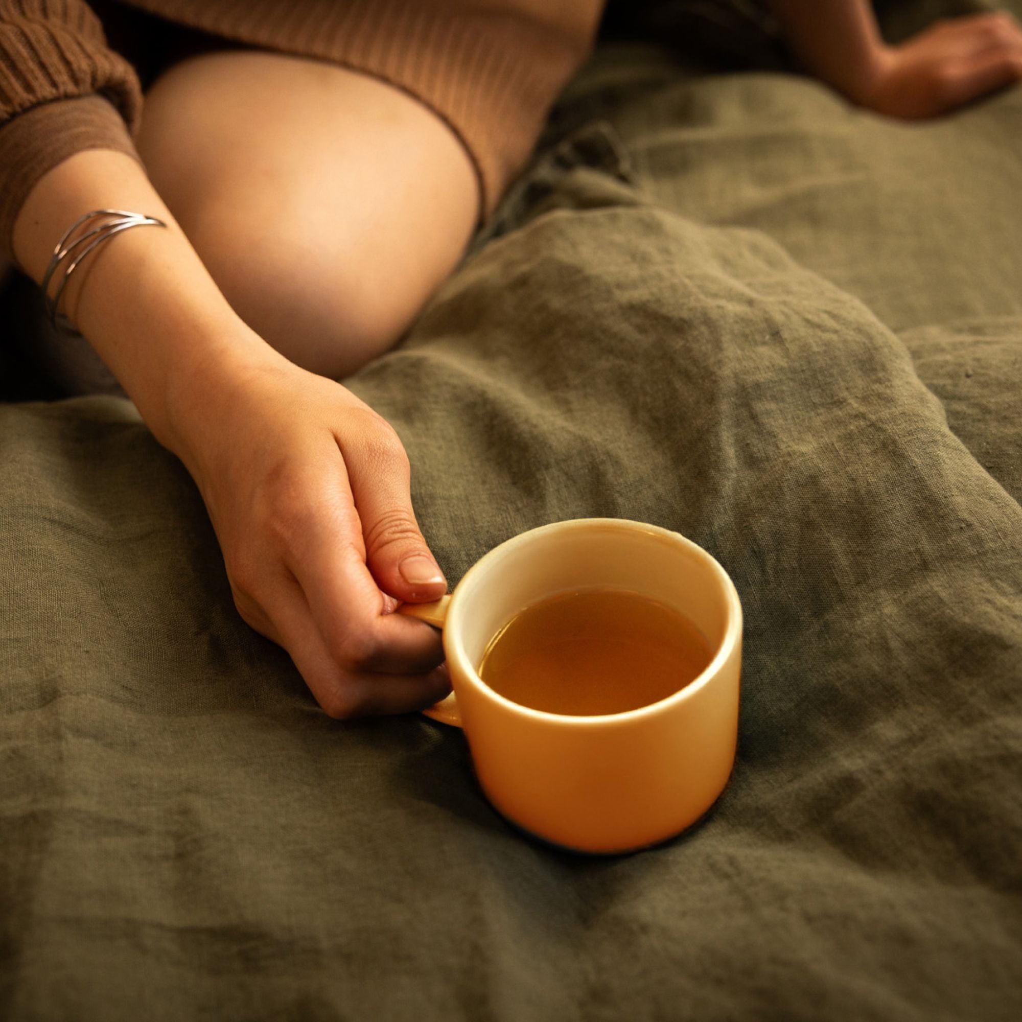 Person hält gelbe Keramiktasse mit Tee vor olivgrünem Hintergrund, trägt braunen Pullover und Silberarmband.
