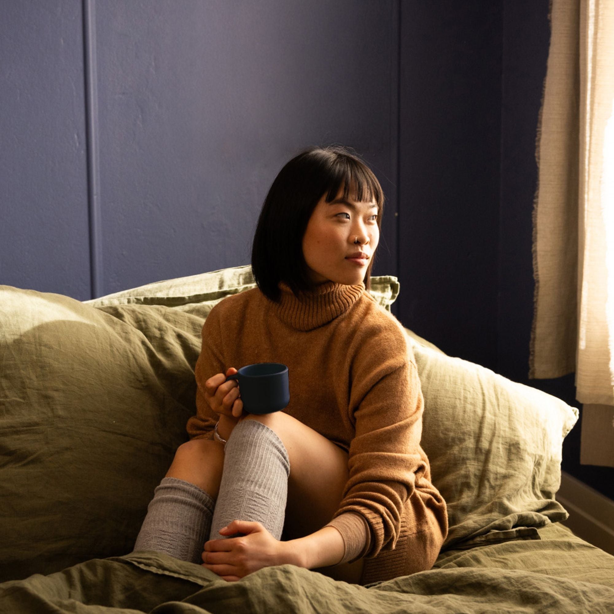Junge Frau in braunem Rollkragenpullover und grauen Kniestrümpfen mit Tasse auf olivgrünem Bett im gemütlichen Raum.