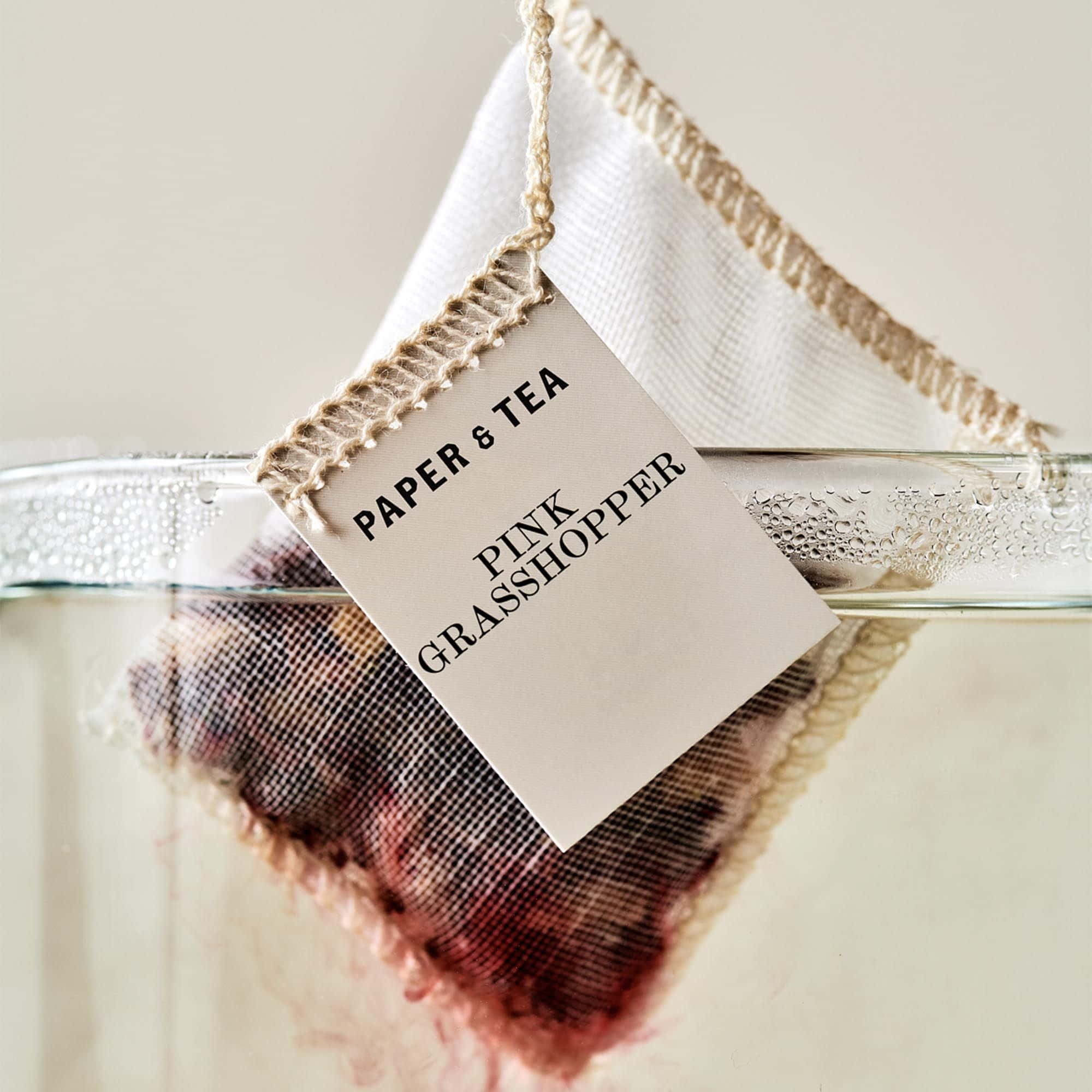 Nahaufnahme eines Papier & Tea Teebeutels 'Pink Grasshopper' in klarem Wasser mit rosa Blüten.