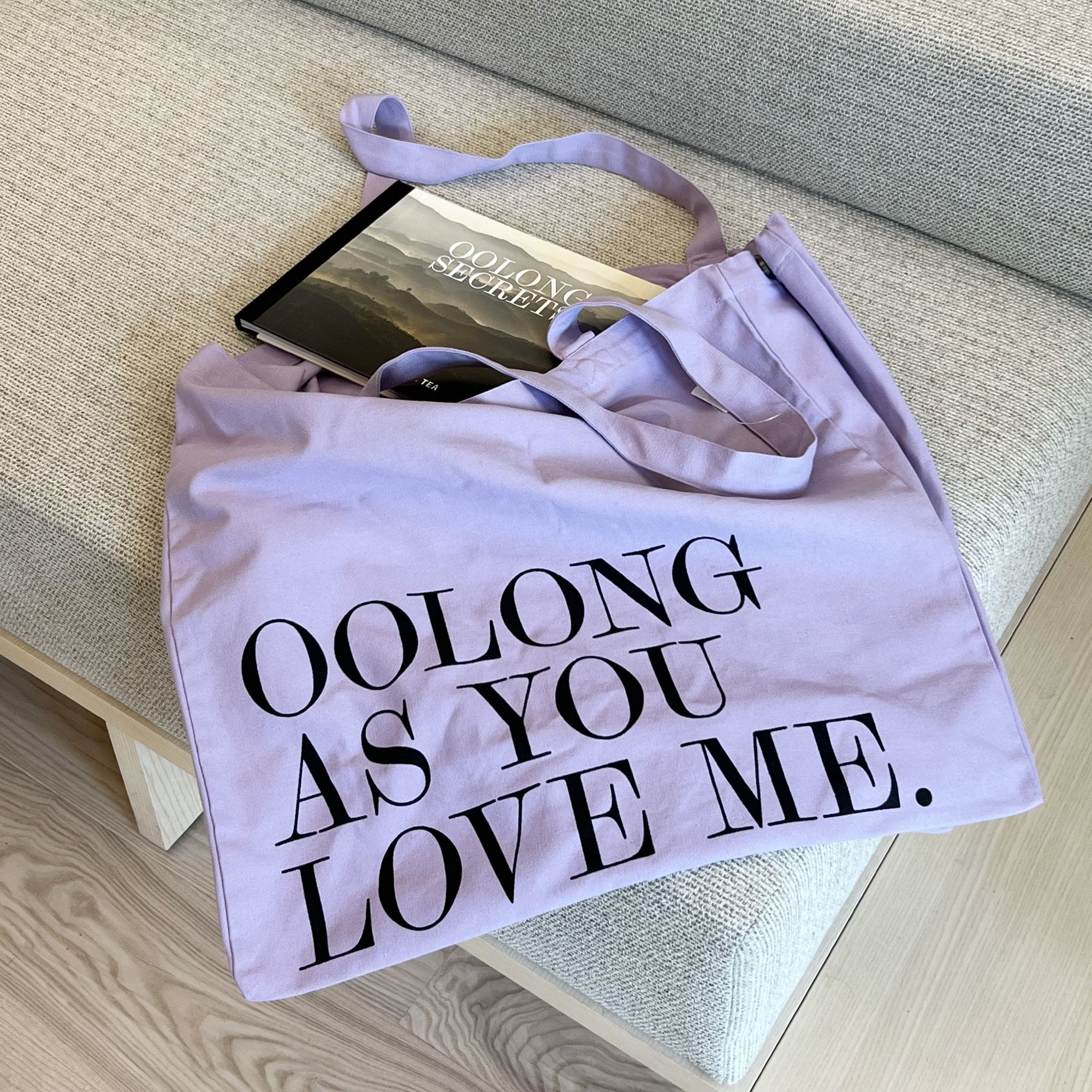 Hellvioletter Stoffbeutel mit Aufschrift 'OOLONG AS YOU LOVE ME' neben Buch 'OOLONG SECRET TEA' auf Sitz.