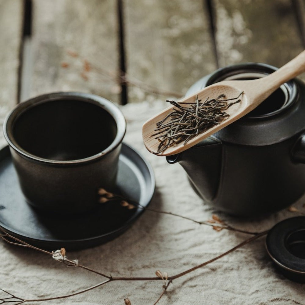 Schwarze Keramiktasse auf Unterteller, daneben schwarze Teekanne mit Holzlöffel und losem Tee, vor Holzhintergrund.
