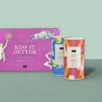Rosa KISS IT BETTER Geschenkbox mit weißen Details neben bunten Teedosen auf grünem Hintergrund.