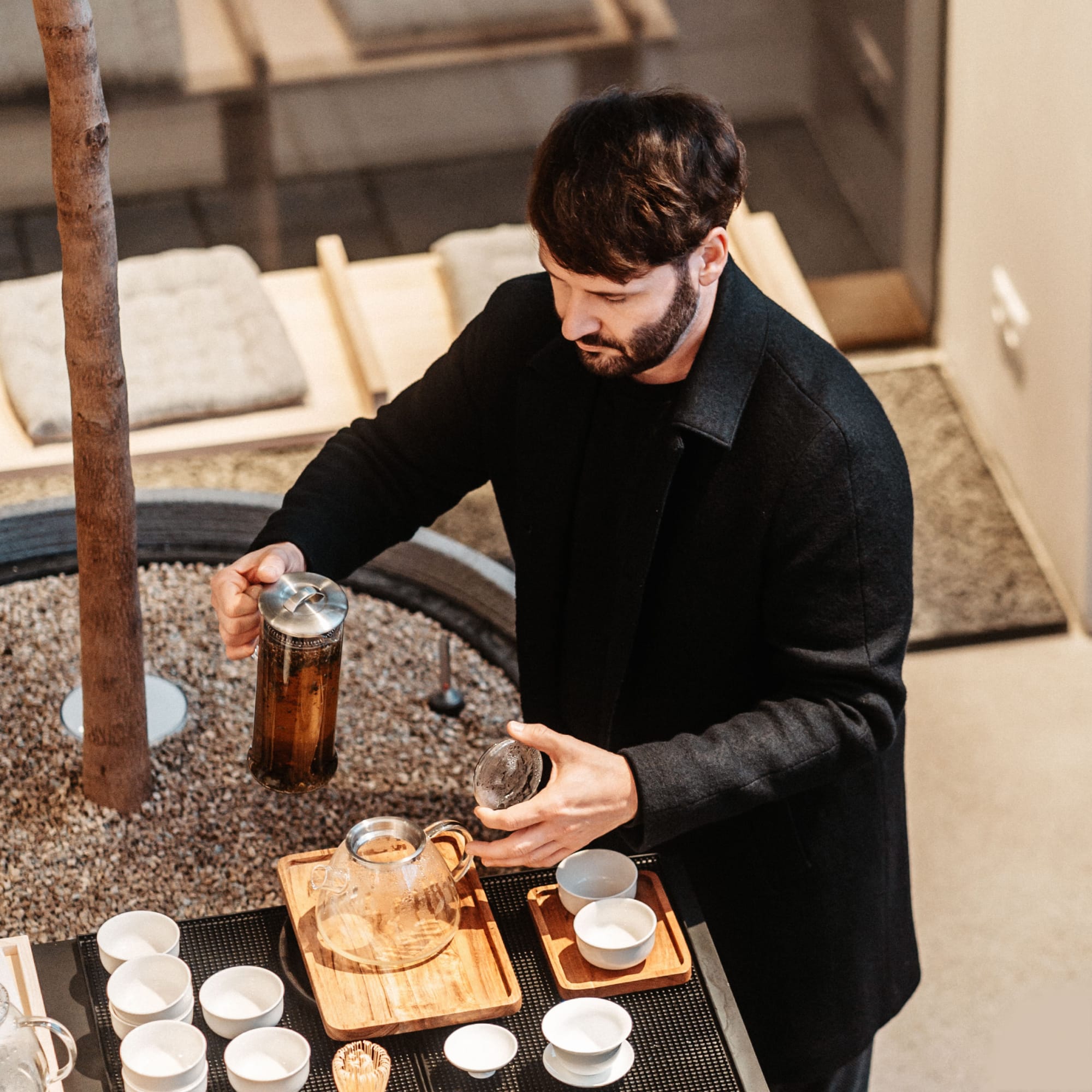 Mann in schwarzem Mantel gießt Tee aus Glaskanne an Tresen mit weißen Tassen in moderner Teestube