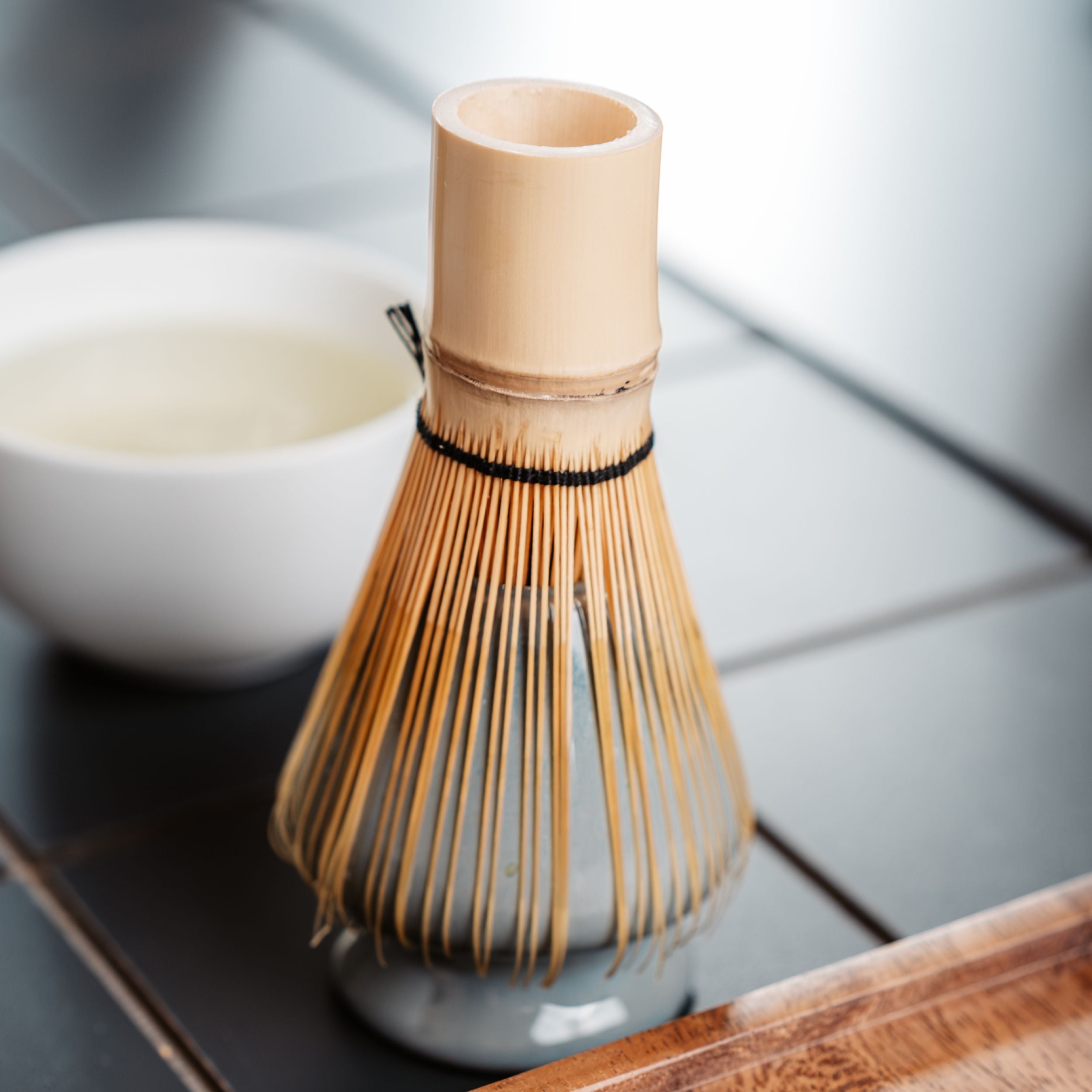Traditioneller Bambus-Matcha-Teebesen auf dunkler Fliese, weiße Schale mit Matcha-Tee im Hintergrund.
