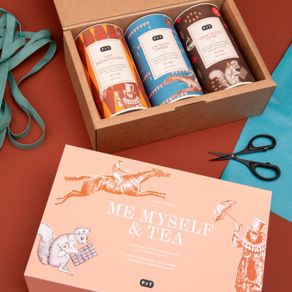 Elegantes Paper & Tea Geschenkset mit farbigen Teedosen, Pfirsich-Deckeln und grünem Band auf braunem Hintergrund.