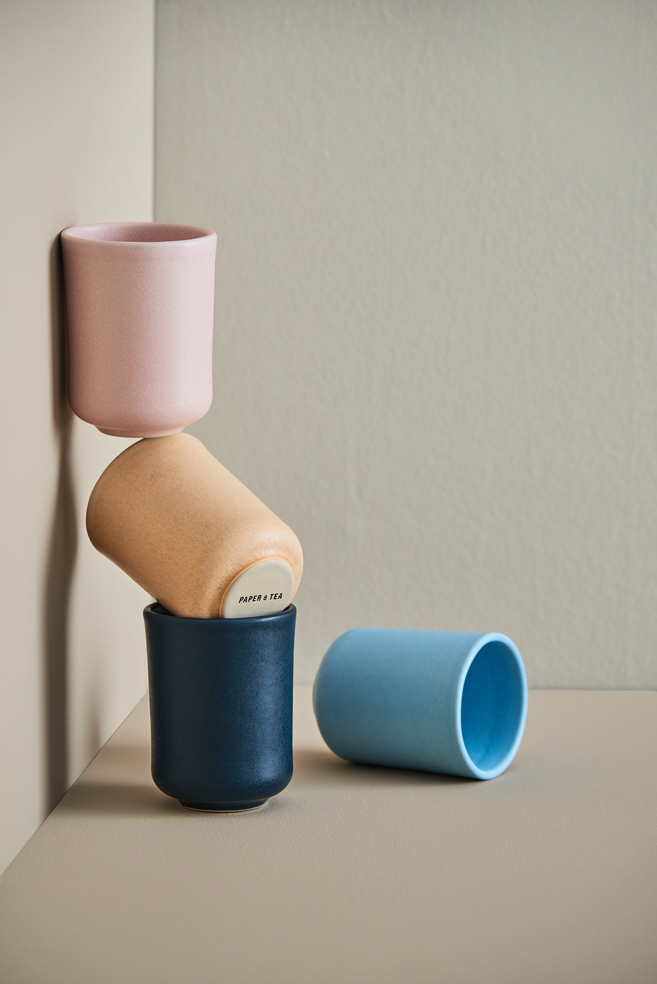 Drei gestapelte Keramiktassen in Pastellfarben mit PAPER & TEA Aufschrift auf der mittleren Tasse, neben einer liegenden hellblauen Tasse auf beigem Untergrund.