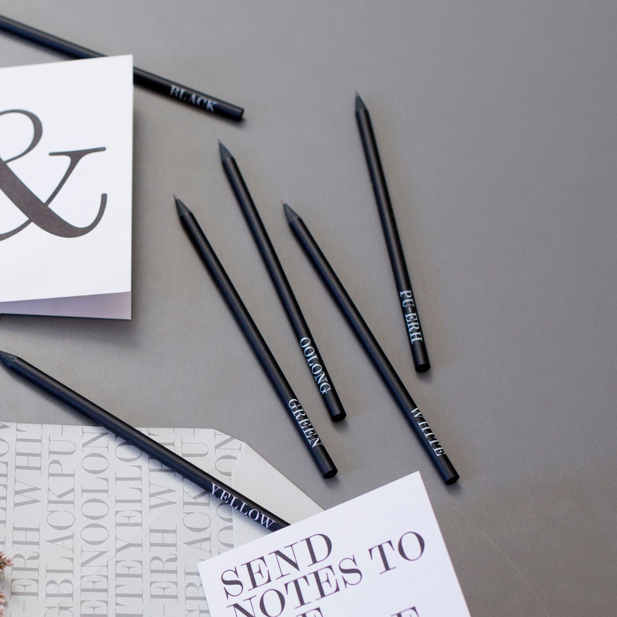 Sechs schwarze Bleistifte mit Teesorten-Beschriftungen auf grauem Hintergrund neben beschrifteten Zetteln.