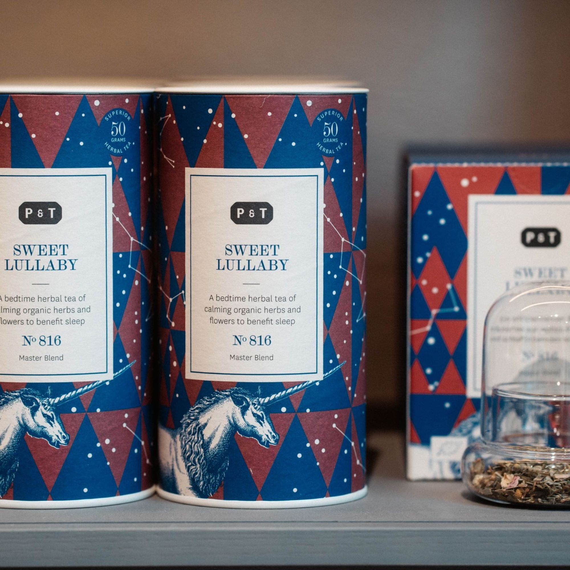 Zwei dekorative Teedosen und eine Box P & T Sweet Lullaby Kräutertee, umgeben von losem Tee in Glasbehälter.