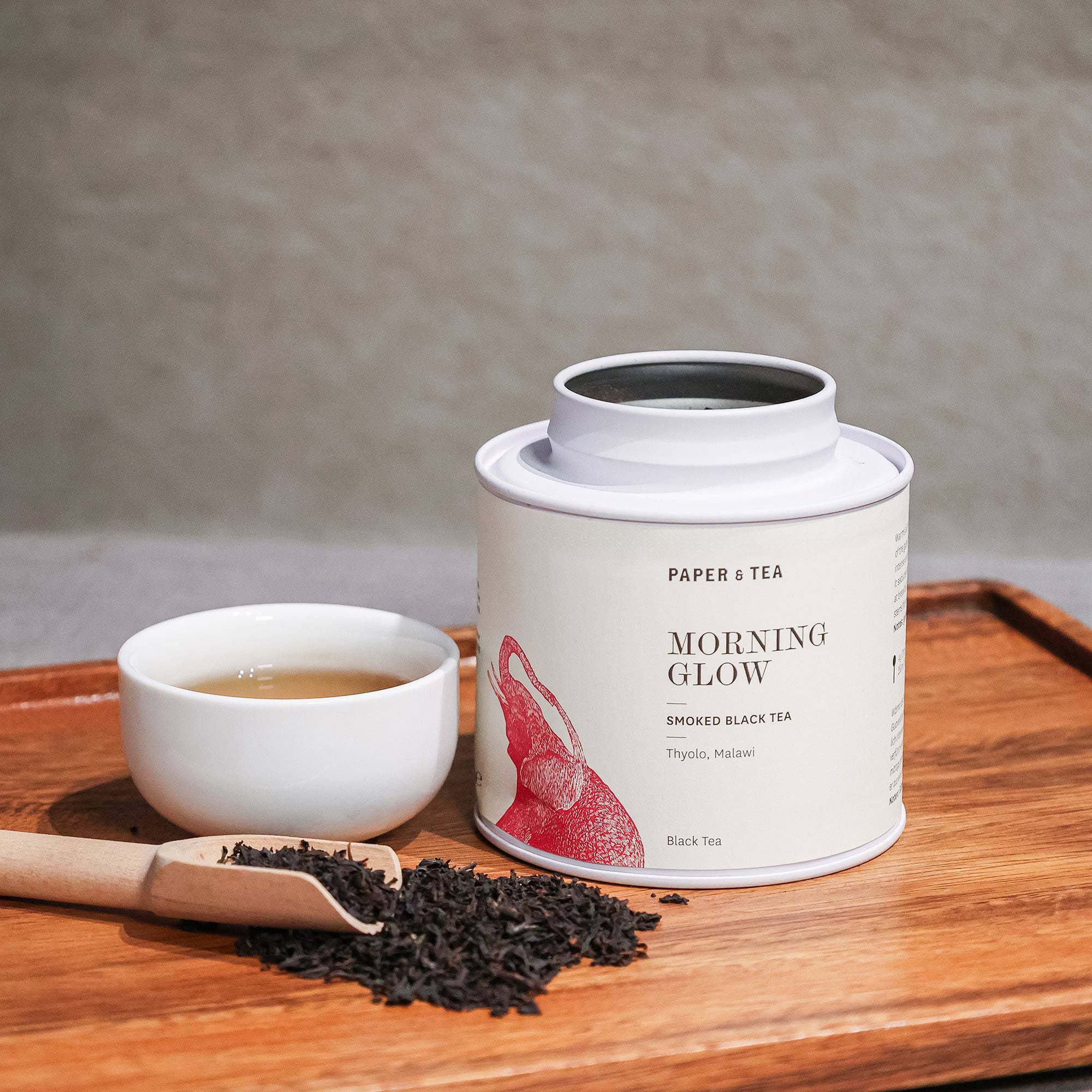 Weißer Teebehälter 'Morning Glow' Schwarztee, Keramiktasse mit Tee, Holzlöffel auf Holztablett.