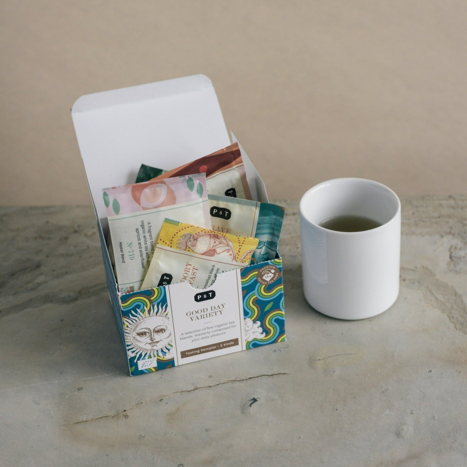Offene Tee-Probierbox „P & T GOOD DAY VARIETY“ mit Bio-Teesorten und weiße Tasse auf Steinuntergrund.