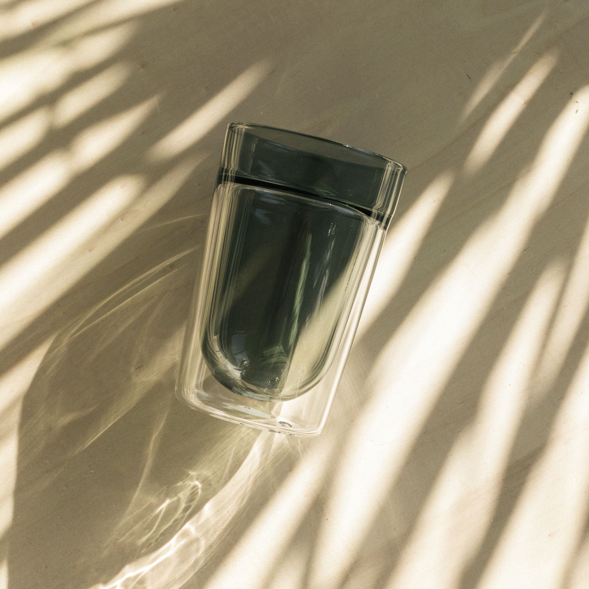 Transparenter doppelwandiger Glasbecher auf hellem Untergrund mit Palmblatt-Schatten im warmen Licht.