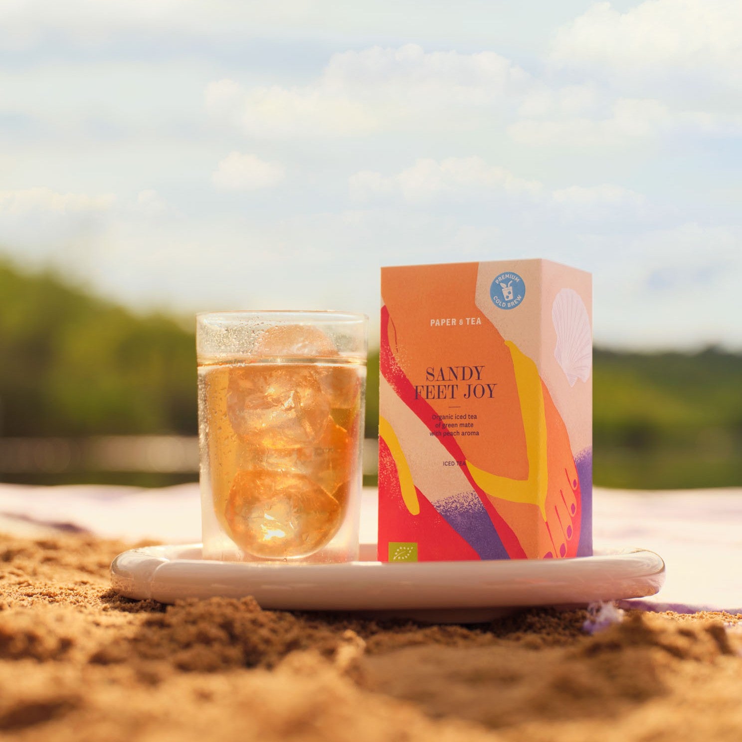 Ein Glas ICE-Tee 'Sandy Feet Joy' von PAPER & TEA neben bunter Verpackung auf einem weißen Teller im Sand.
