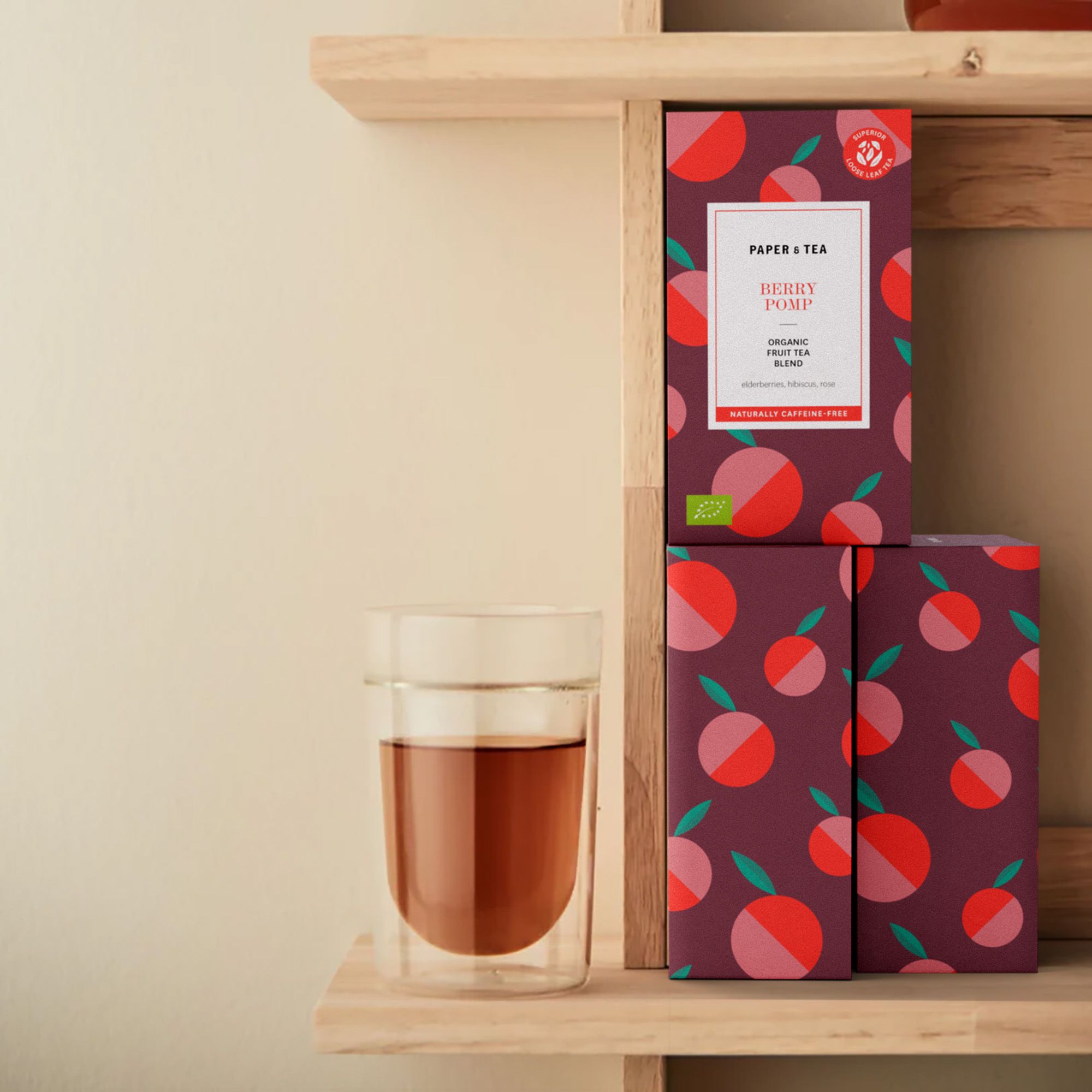 Drei stapelbare Beutel Paper & Tea Berry Pomp Früchtetee neben Glas mit rotem Tee auf Holzregal.