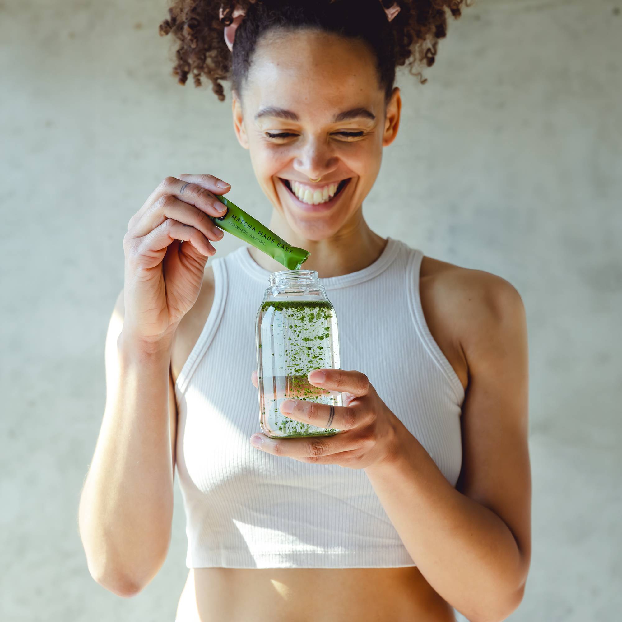 Junge Frau in weißem Crop-Top gießt grünes Matcha-Pulver aus P&T Sachet in Glasflasche mit Wasser ein.