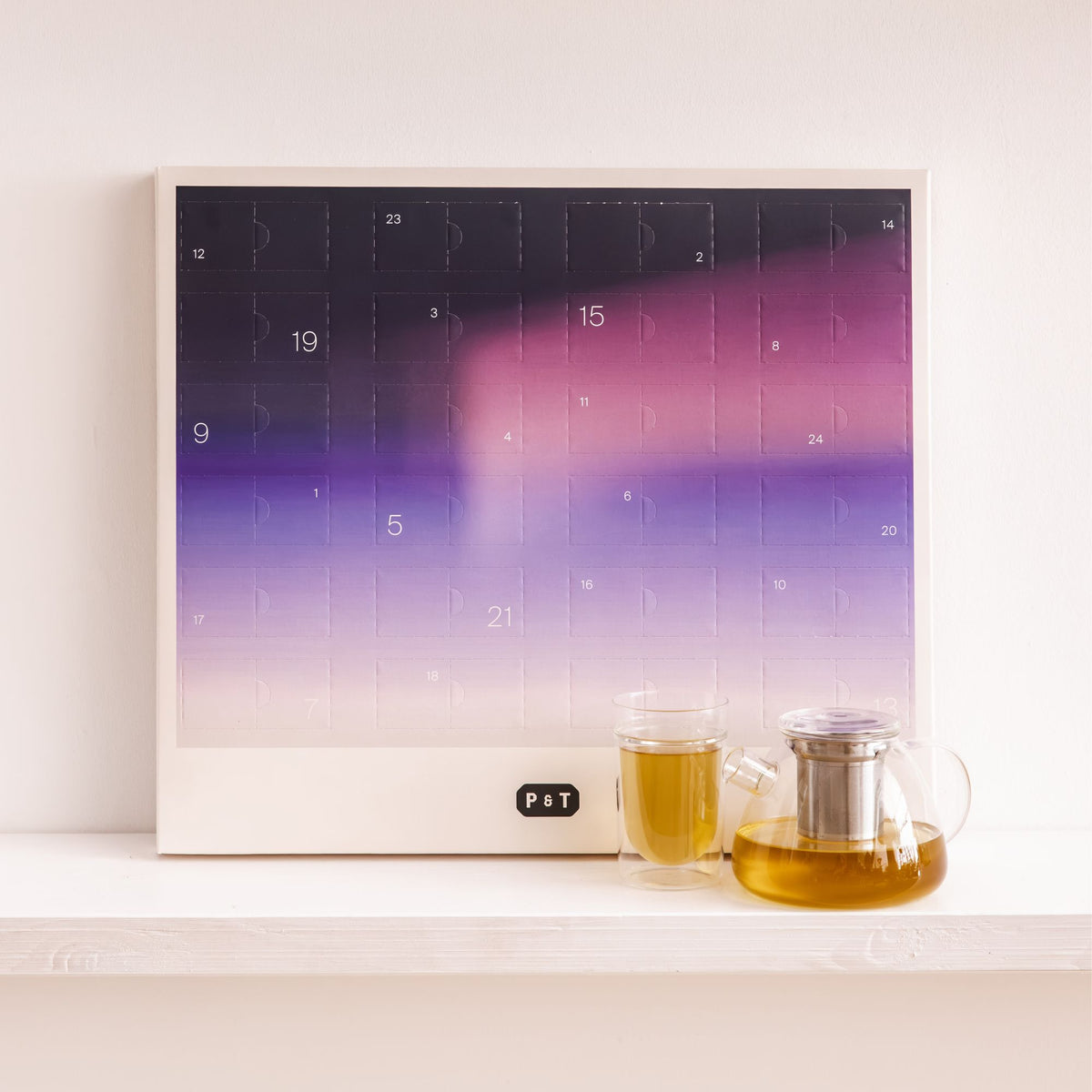 Minimalistischer Adventskalender von P&T mit lila-cremem Farbverlauf, nummerierten Türen und Teekanne sowie Teeglas auf weißem Regal.
