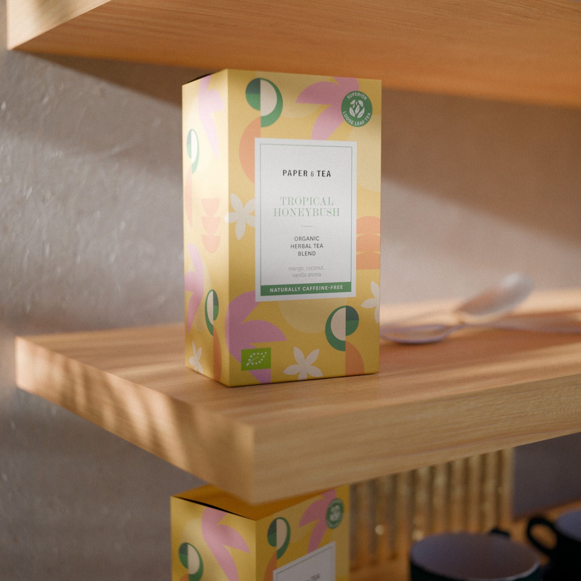 Gelbe Verpackung des Tropical Honeybush Kräutertees von Paper & Tea auf Holzregal.