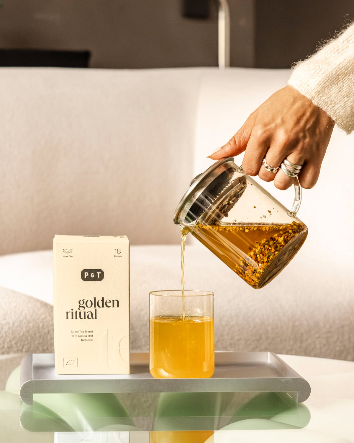 Hand gießt goldgelben Gewürztee von P&T Golden Ritual in Glasbecher auf silbernem Tablett.