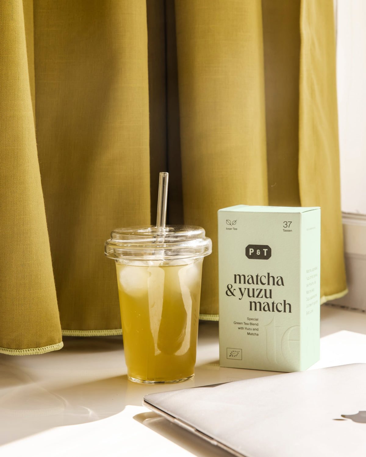 Transparenter Kunststoffbecher mit eisgekühltem Matcha & Yuzu Grüntee neben pastellgrüner P&T Teeschachtel.