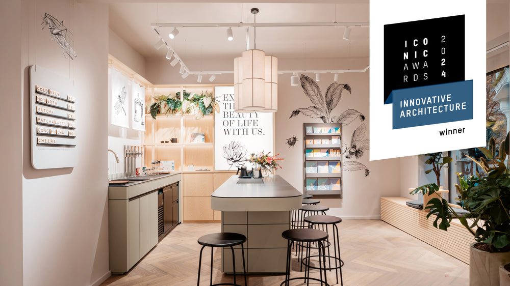 Minimalistisches Café mit Insel, sechs schwarzen Hockern, Pflanzen und Tafel mit Teeangebot.