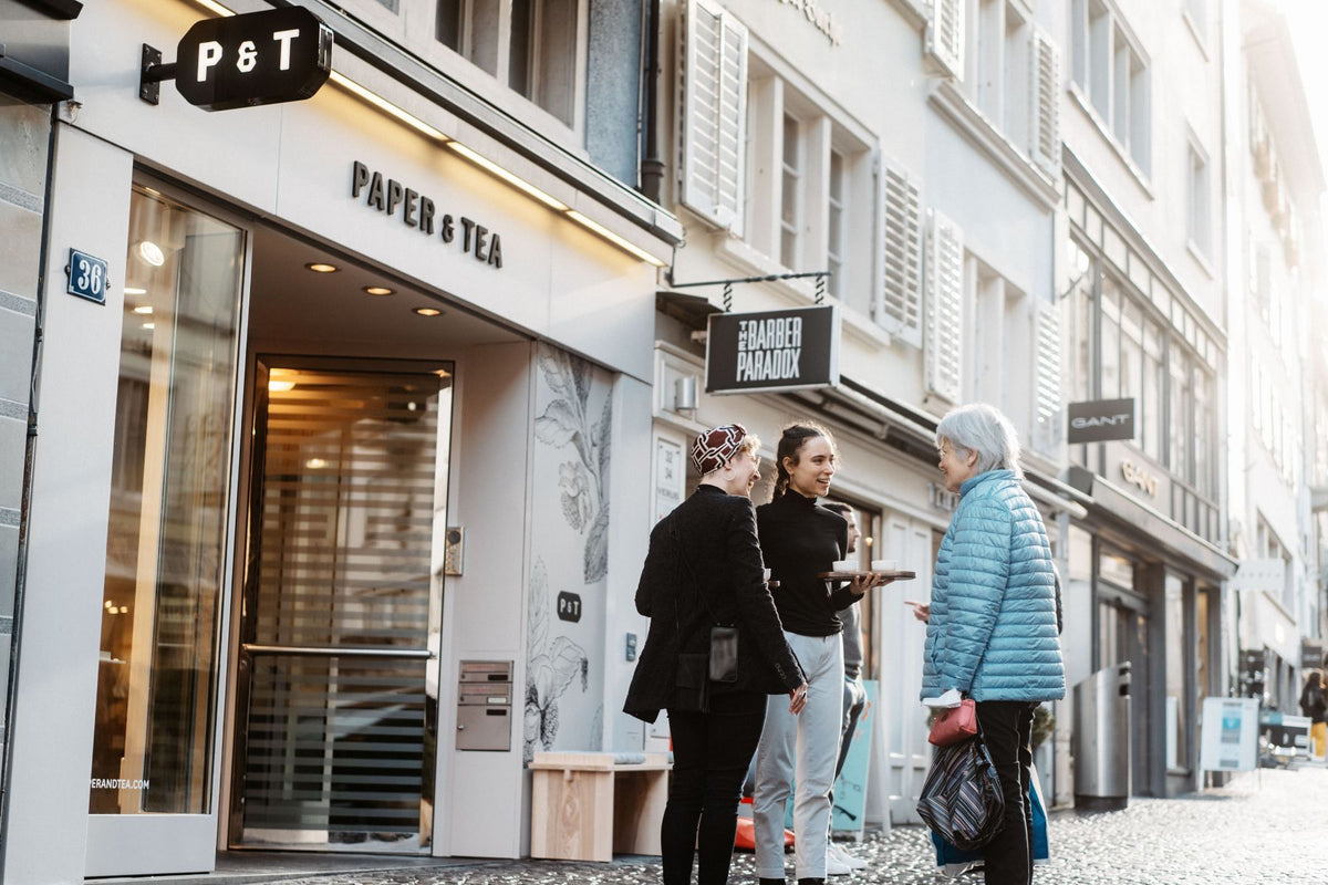 Belebter Straßenabschnitt mit dem Laden Paper & Tea, leuchtendem P & T Schild, Personen mit Teetassen