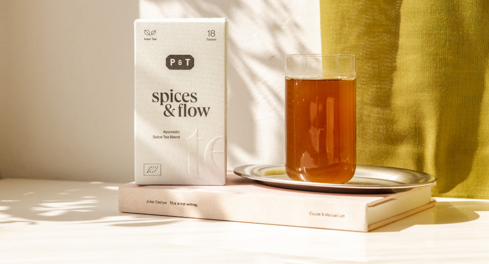 Weiße P&T Ayurvedische Gewürz-Teemischung auf Buch, Glas Tee auf Metalltablett vor gelbgrünem Stoff